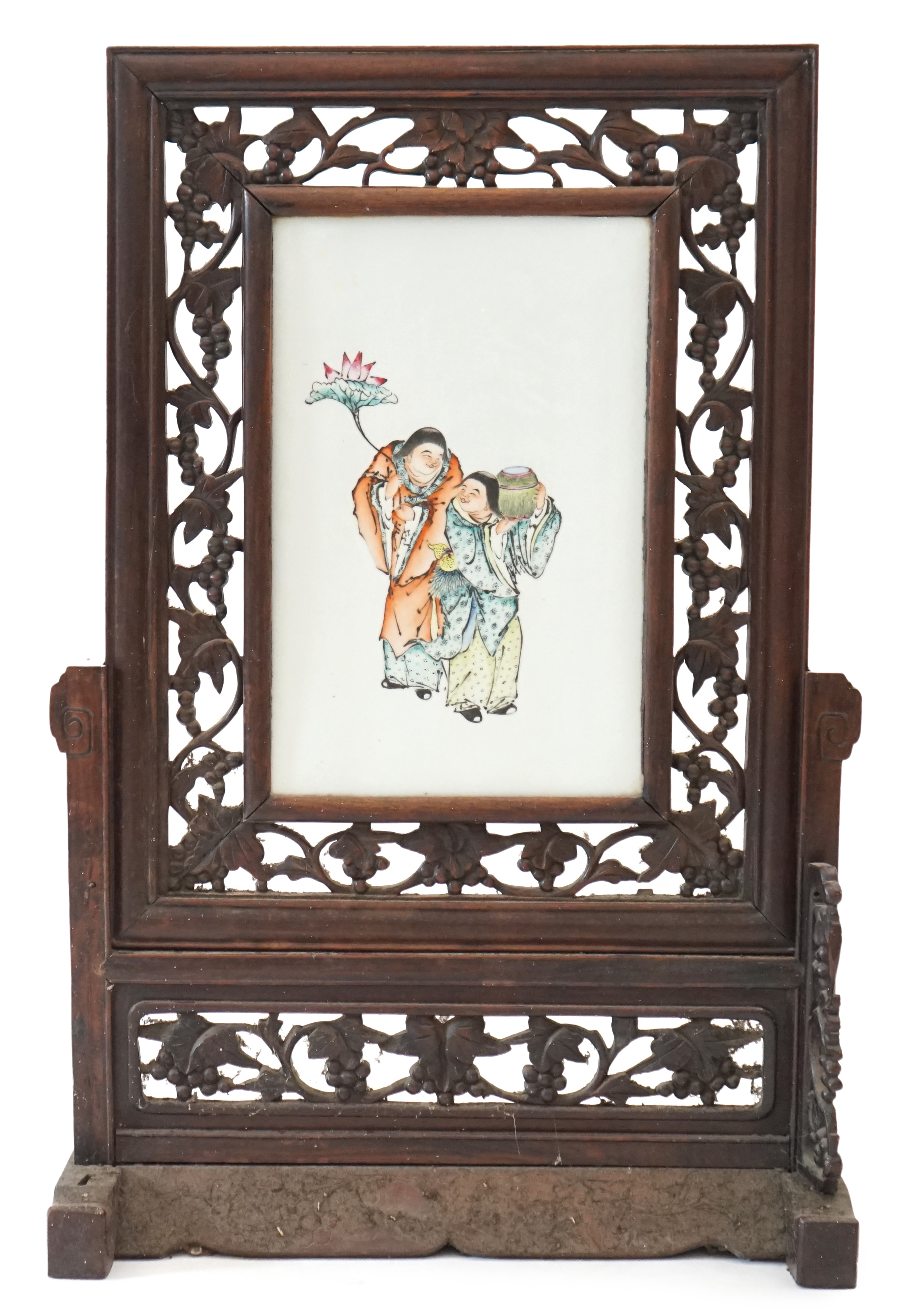 A Chinese enamelled porcelain and hardwood table screen, Republic period, painted with the HeHe Erxian, holding a sprig of lotus and a box and cover, the plaque 37.5cm x 24cm, the wood stand and frame carved with vines,